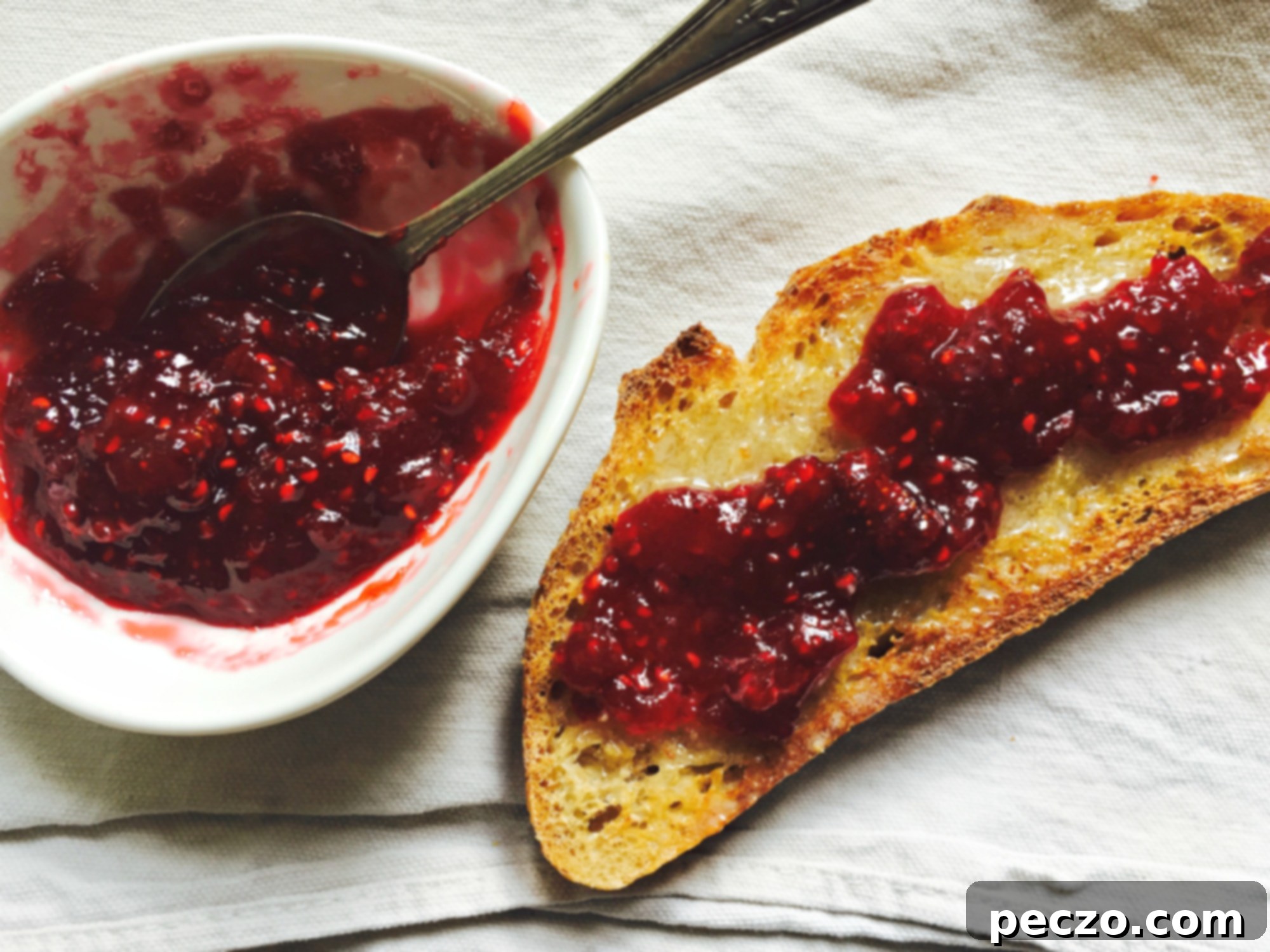 A vibrant jar of homemade red berry jam sits beside a slice of rustic bread, perfectly toasted and ready to be spread with deliciousness. The rich color of the jam hints at its fresh, fruity flavor, inviting you to savor a simple pleasure.