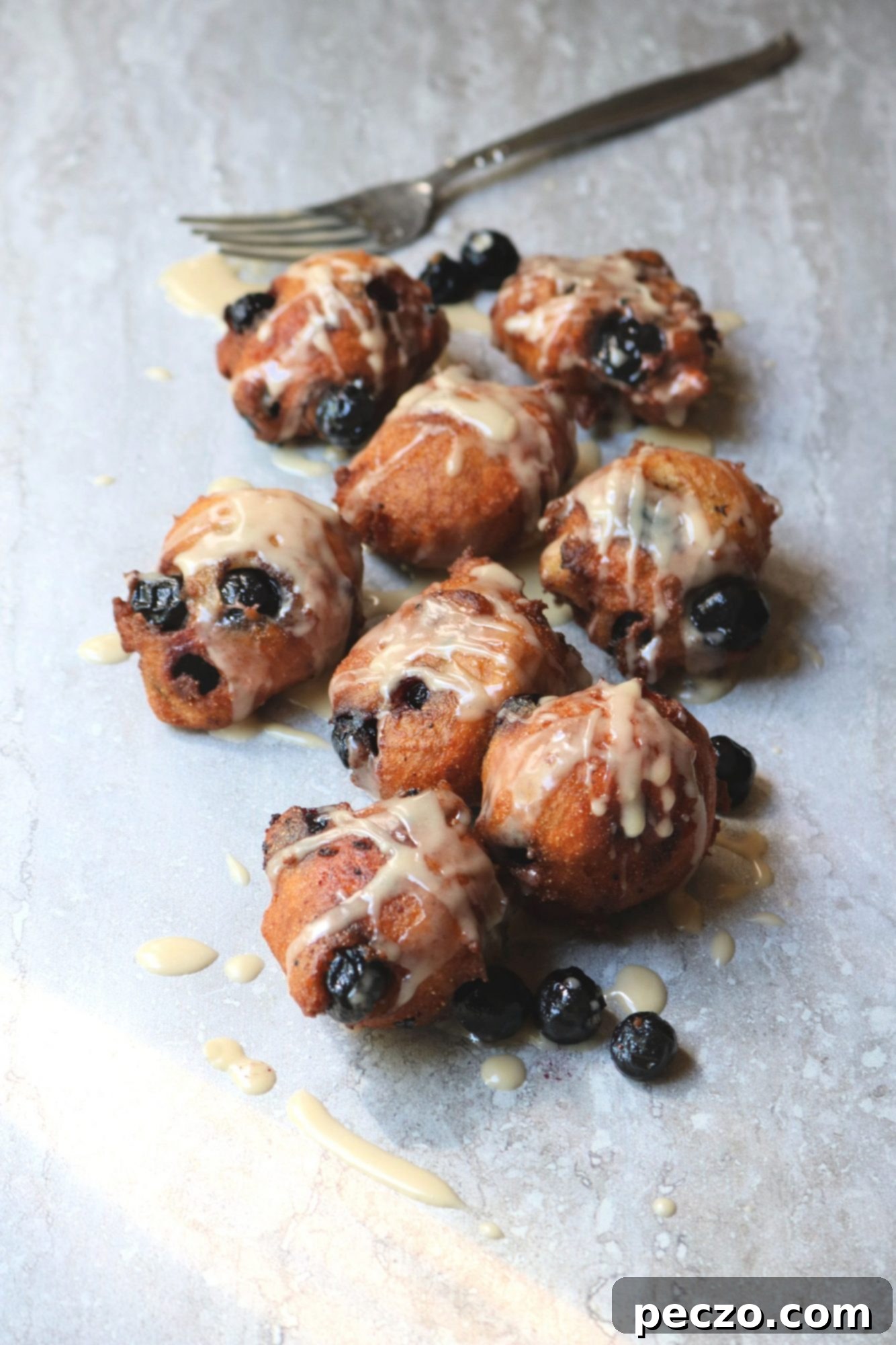 A close-up of a stack of blueberry corn fritters on a white plate, generously drizzled with maple glaze, with fresh blueberries and maple syrup in the background.