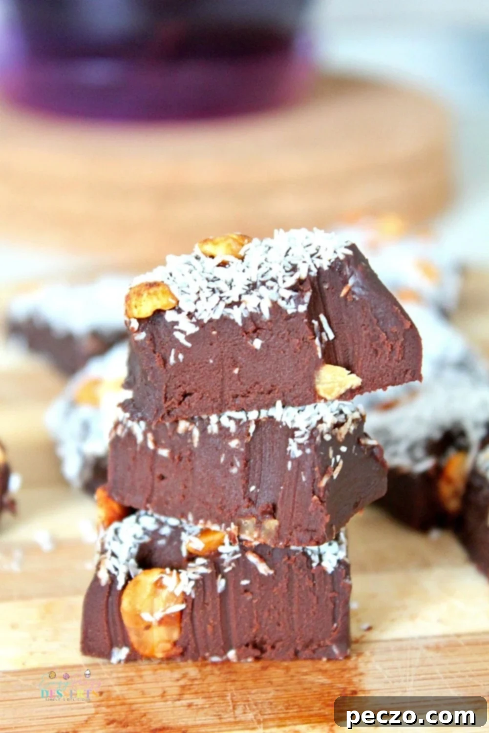 Three decadent pieces of 2-ingredient chocolate fudge, garnished with coconut flakes and chopped nuts, resting on a rustic wooden surface